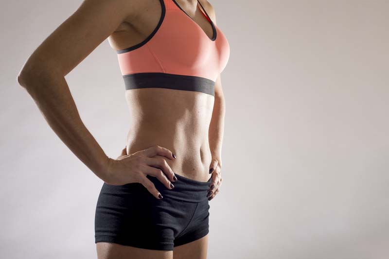 Cropped close up body of fit woman wearing shorts and sport top showing slim beautiful stomach and abs in diet fitness and healthy lifestyle concept on grey background